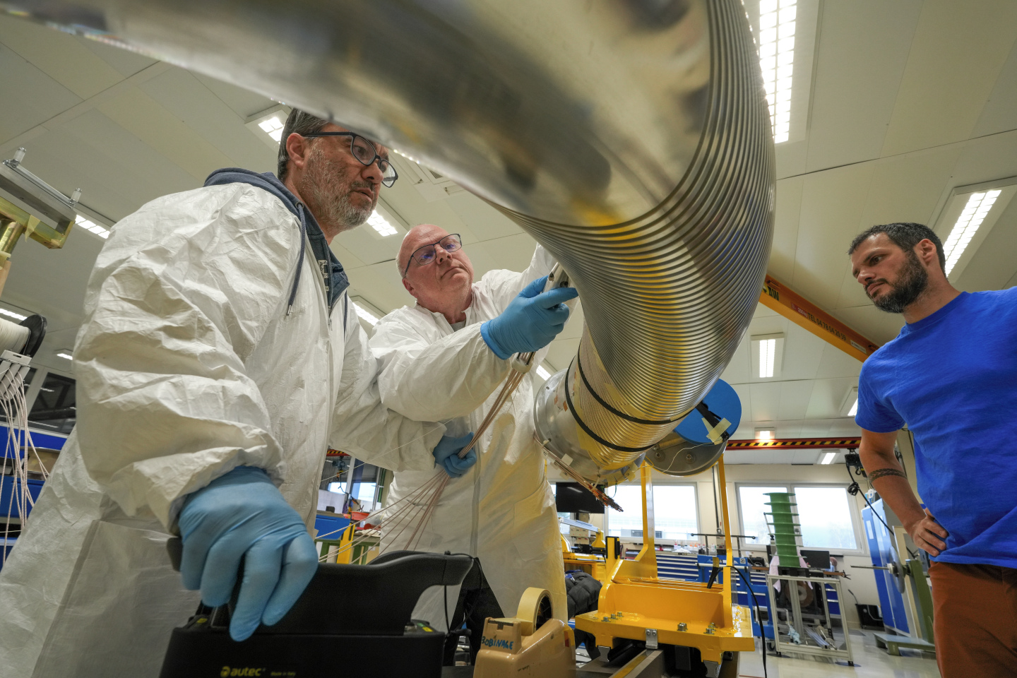 Two men in protective suits guide a bundle of ropes made from wires into the grooves of a large, reflective metal former, while a third person observes the process nearby